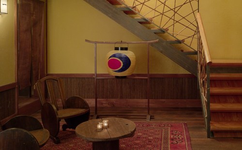 A cozy interior with a decorative lantern, wooden chairs, a small table with candles, and a staircase in the background.