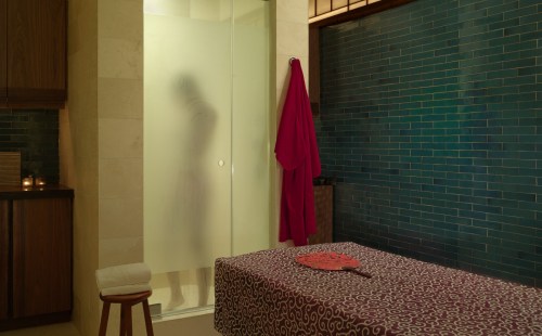 A serene bathroom scene with a shower, a red robe hanging, and a massage table adorned with a patterned cloth.
