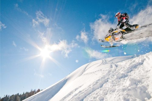 A snowmobiler jumps off a snowy hill under a bright blue sky with the sun shining. Snow sprays behind them.