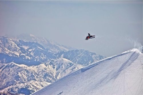 A snowmobiler jumps off a snowy peak with mountains in the background under a clear sky.