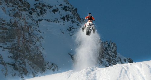 A person on an ATV jumps over a snowy slope, with mountains in the background and snow flying around.
