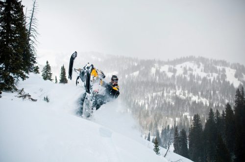 A snowmobiler performs a wheelie in deep snow, surrounded by a snowy mountain landscape and evergreen trees.