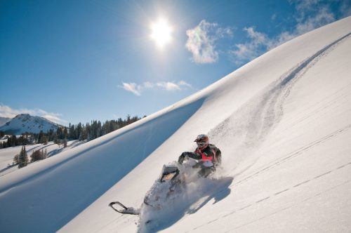 A snowmobiler speeds down a snowy slope under a bright sun, surrounded by trees and mountains.