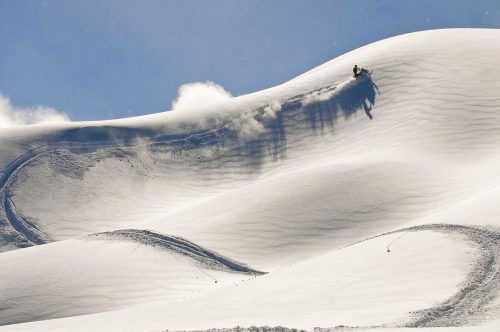 A snowmobiler rides down a snowy dune under a clear blue sky, leaving tracks in the pristine snow.