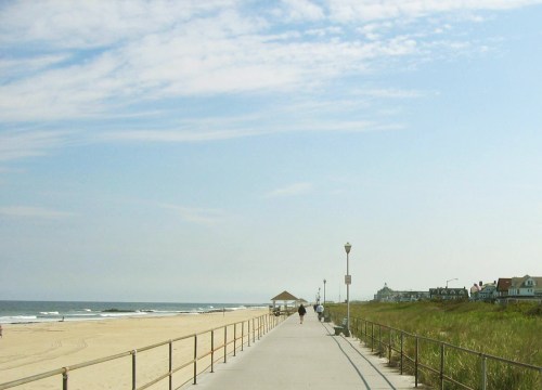 A scenic beach boardwalk with a view of the ocean, sandy shore, and distant houses under a blue sky.