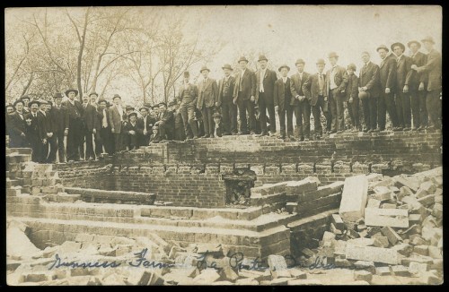A large group of men in suits stands on a brick foundation, surrounded by rubble and trees, in a vintage photograph.