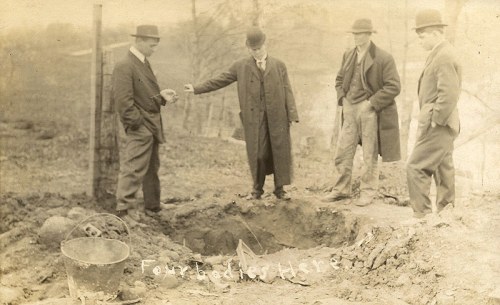 Four men in hats stand around a large hole in the ground, examining something with serious expressions.