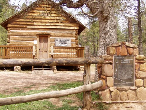 Historic log cabin with a plaque in front, surrounded by trees and a wooden fence.
