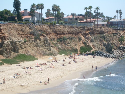 A sunny beach scene with people sunbathing and swimming, surrounded by cliffs and palm trees.