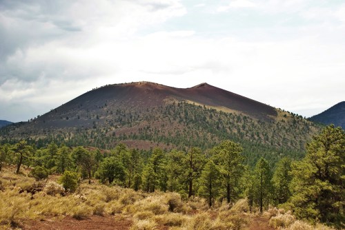 A volcanic mountain rises above a forest of green trees under a cloudy sky.