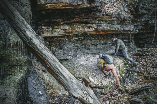 Two hikers exploring a rocky area with a cliff, surrounded by fallen leaves and trees.