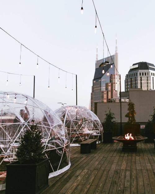 Rooftop view with transparent domes, string lights, and a fire pit, set against a city skyline at dusk.