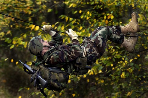 A soldier in camouflage gear rappels down a rope amidst autumn foliage.