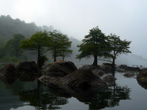 Misty landscape with trees on rocky outcrops reflected in calm water, surrounded by a foggy atmosphere.