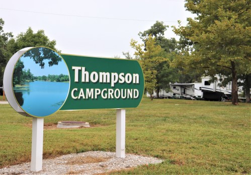 Sign for Thompson Campground featuring a scenic lake view, surrounded by trees and RVs in the background.