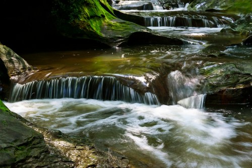 A serene stream flows over moss-covered rocks, creating small waterfalls in a lush, green forest setting.