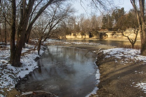 A serene winter scene featuring a partially frozen river, snow-covered banks, and bare trees along the shoreline.