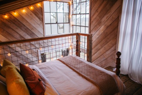 Cozy bedroom with a wooden interior, large windows, and warm string lights, featuring a neatly made bed.