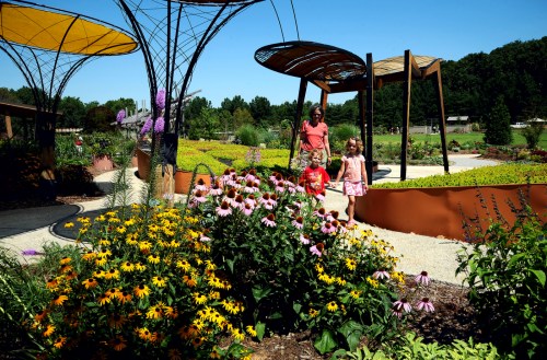 A woman and two children walk through a vibrant garden filled with colorful flowers and unique structures on a sunny day.
