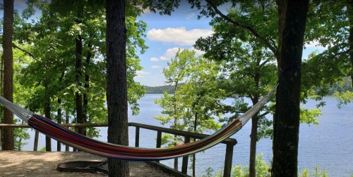 A colorful hammock hangs between trees, overlooking a serene lake and blue sky with fluffy clouds.