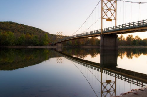 A yellow suspension bridge spans a calm river, reflecting the surrounding trees and hills at sunset.
