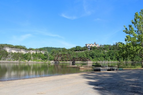 A serene river scene with a bridge, sandy shore, and a house on a hill surrounded by lush greenery under a clear blue sky.