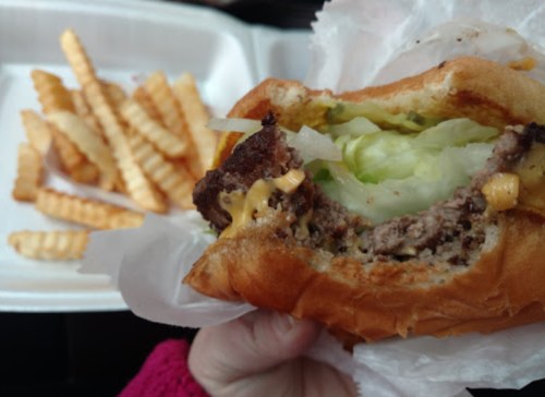A hand holding a half-eaten cheeseburger with lettuce, next to a container of crinkle-cut fries.