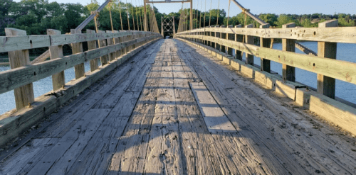 A wooden bridge stretches over water, with sunlight casting shadows on the weathered planks and surrounding greenery.