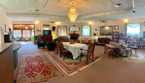 A spacious, well-furnished living room with a chandelier, dining table, and cozy seating areas, featuring bookshelves and artwork.