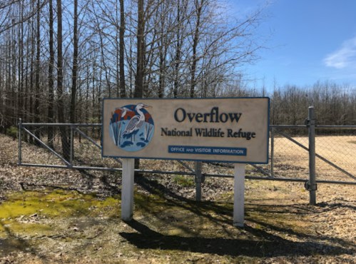 Sign for Overflow National Wildlife Refuge, featuring a heron graphic, with trees and a field in the background.