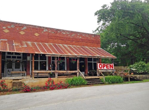 A rustic storefront with a metal roof, featuring an "OPEN" sign and surrounded by greenery and flowers.