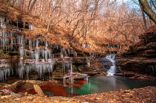 A serene waterfall surrounded by rocky cliffs, icicles, and autumn foliage under a clear blue sky.