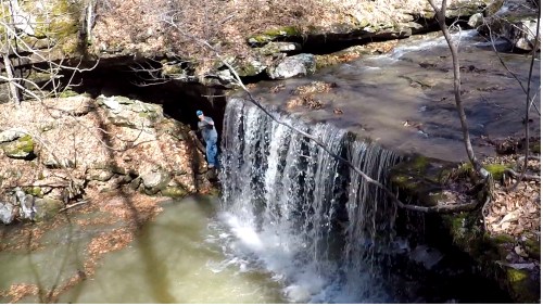 A person stands near a waterfall cascading over rocky terrain, surrounded by trees and fallen leaves.