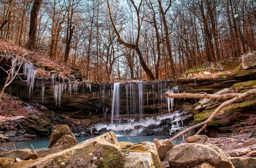 A serene waterfall cascades over rocky ledges, surrounded by trees and icicles in a tranquil forest setting.