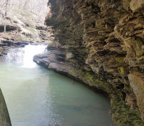 A serene river flows between rocky cliffs, with a small waterfall in the background and greenery along the banks.