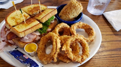 A plate with a sandwich, onion rings, a biscuit, and a side of mustard on a wooden table.