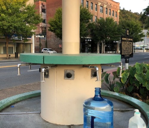 A water fountain with multiple spouts, surrounded by greenery and buildings in the background. A water jug is nearby.