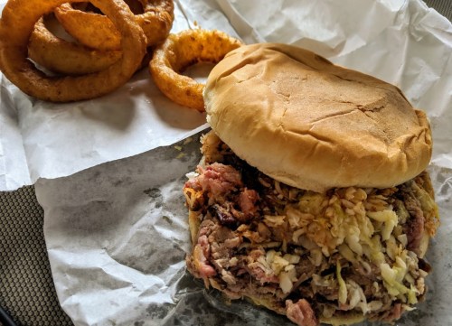 A pulled pork sandwich topped with coleslaw, served with crispy onion rings on the side.