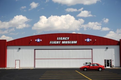 Exterior of the Legacy Flight Museum, featuring a red and white building with a blue sky and clouds above.
