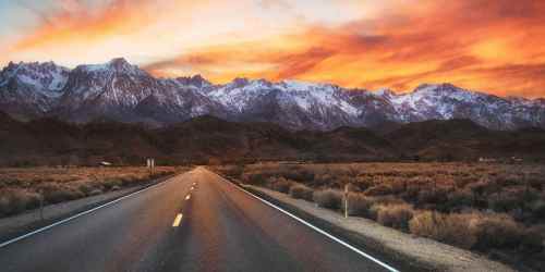 A scenic road leads towards snow-capped mountains under a vibrant sunset sky.