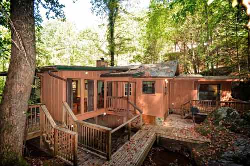 A rustic wooden house surrounded by trees, featuring a deck and a pathway leading to the entrance.