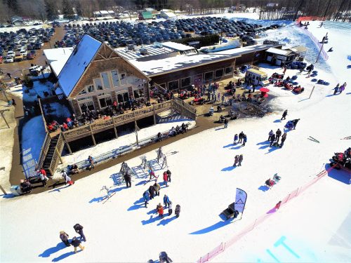 A snowy landscape with a busy ski lodge, people enjoying outdoor activities, and a large parking lot in the background.