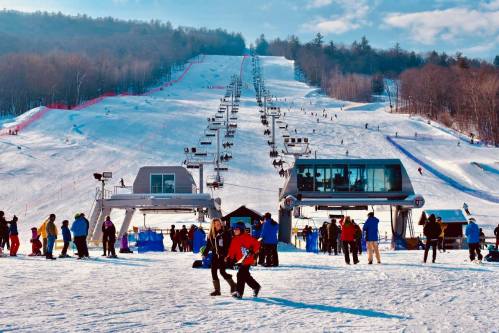 Ski resort scene with people on a snowy slope, ski lifts in the background, and a clear blue sky.