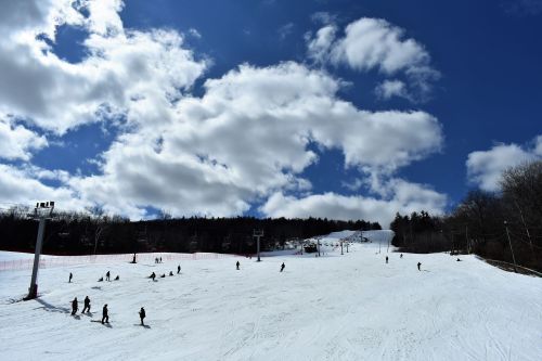 Skiers and snowboarders on a snowy slope under a blue sky with fluffy clouds and ski lifts in the background.