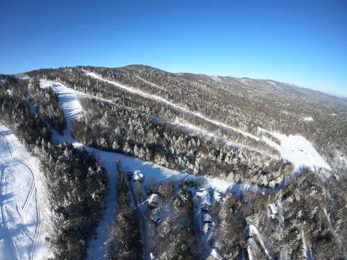 Aerial view of snow-covered mountains and trees under a clear blue sky, with ski trails visible in the landscape.