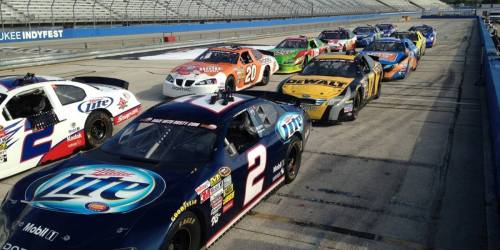 A lineup of colorful NASCAR race cars parked on a track, with empty grandstands in the background.