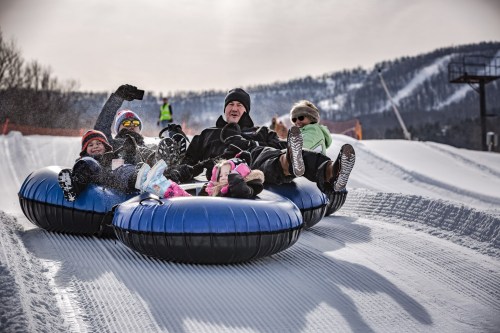 A family enjoys snow tubing together, laughing and having fun on a snowy slope with mountains in the background.