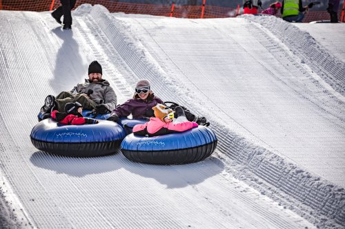 Two people sit on inflatable tubes at the top of a snowy hill, ready to slide down. Bright winter clothing and a fun atmosphere.