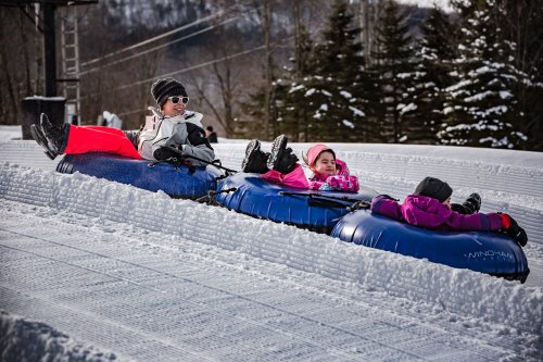 Two children in colorful winter gear enjoy tubing down a snowy slope on inflatable tubes.