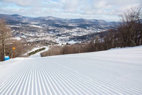 A snowy slope with groomed lines, overlooking a valley and mountains under a partly cloudy sky.